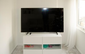 Un televisor de pantalla plana sobre un soporte blanco, un control remoto, un estante de vidrio con algunos objetos y una ventana con cortinas a la derecha.