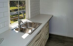 Una cocina con un lavamanos de acero inoxidable, salpicadero de azulejos blancos y ventana con vistas a un jardín.