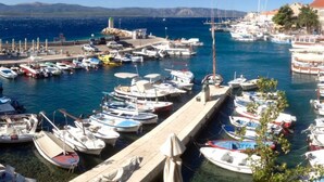 Un puerto deportivo con numerosas embarcaciones amarradas, un muelle con mesas y sillas, y una vista despejada del mar y montañas distantes.