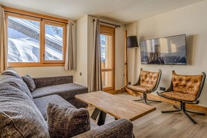 Ein Wohnzimmer mit einer Couch, zwei Sesseln, einem Tisch, einem Fernseher und einem Fenster mit Vorhängen.