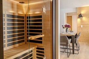 Eine Sauna mit Holzbänken und Glastüren, ein Essbereich mit Tisch und Stühlen sowie eine Holzwand mit einer Leuchte.