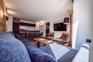 Ein modernes Wohnzimmer mit einer Couch, einem Couchtisch und Sesseln. Es gibt eine Küchenzeile mit Schränken und einem Esstisch. Ein Flachbildfernseher ist an der Wand befestigt.