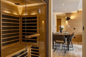 Eine Sauna mit Holzbänken und Glastüren, ein Essbereich mit Tisch und Stühlen sowie eine Holzwand mit einer Leuchte.