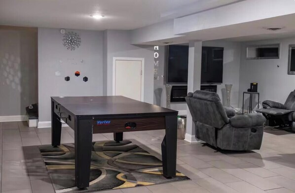 A modern living room with a dark wooden coffee table, two grey reclining chairs, a flat-screen TV mounted on the wall, and a decorative wall piece.