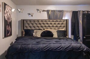 A bedroom with a tufted headboard, a bed with dark bedding, a framed picture on the wall, and butterfly wall decor.