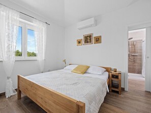A bedroom with a wooden bed, a bedside table, a nightstand, a window with curtains, a wall-mounted air conditioner, and a shower area. - Villa Javor by Interhome (Pula/Krnica)