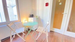 Ein Zimmer mit einem kleinen runden Tisch und zwei Stühlen, einer Tür mit roter Notausgangsangabe und einem Fenster mit Jalousien.