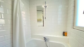 Ein Badezimmer mit weißen Fliesen, einer Badewanne, einer Dusche, einem Spiegel und einem Fenster.