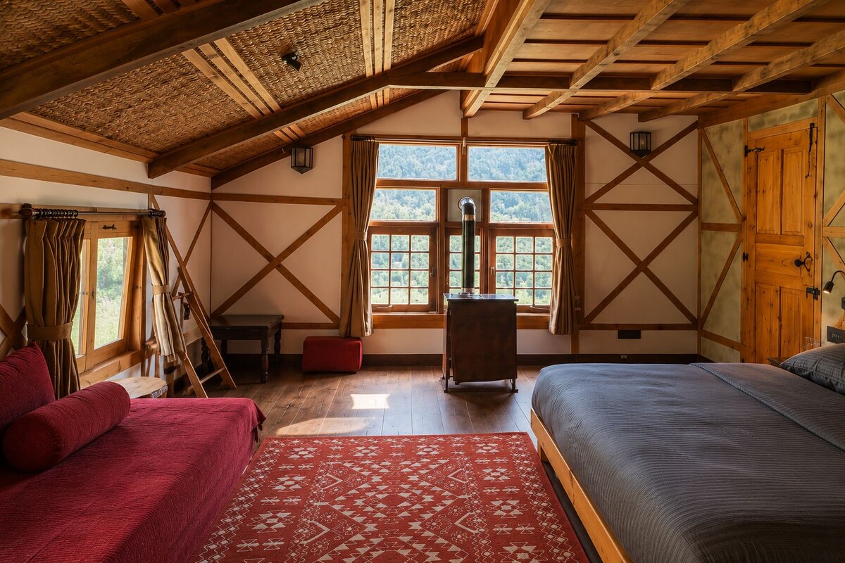 A room with a sloped ceiling, wooden beams, a bed, a red rug, and a window with curtains.