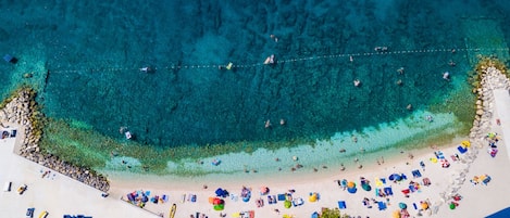 An aerial view of a beach with clear turquoise waters, a sandy shore, and numerous colorful umbrellas and beach chairs.