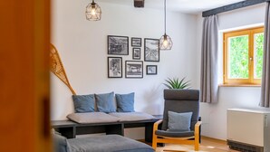 A living room with a sofa, armchair, and a small table. There are framed pictures on the wall and a window with curtains.