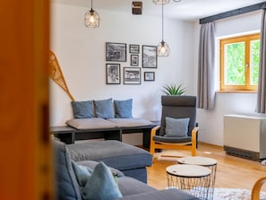 Ein Wohnzimmer mit einer Couch, einem Sessel und einem kleinen Tisch. An den Wänden hängen Bilder und das Fenster ist mit Vorhängen drapiert.