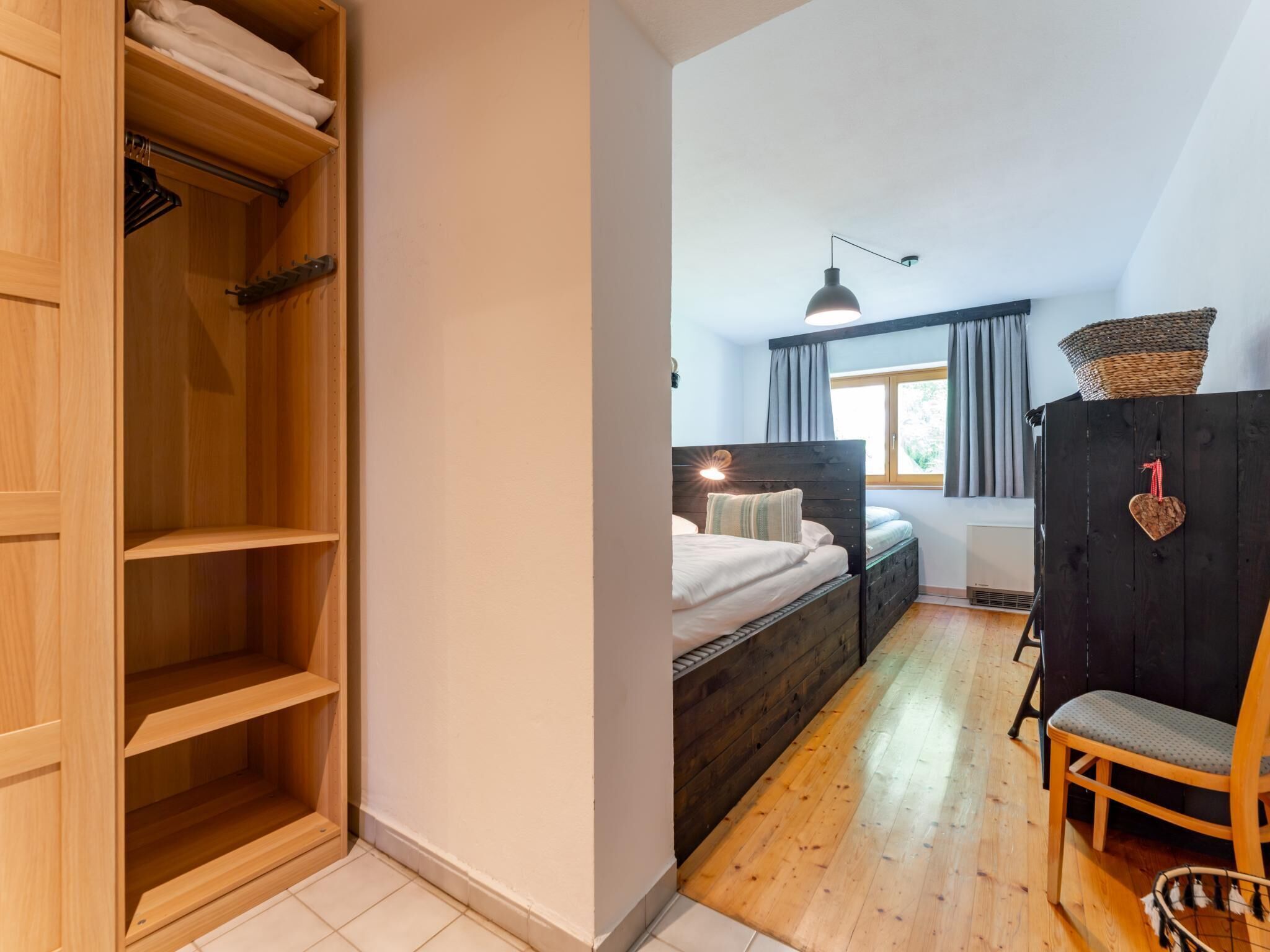 A bedroom with a wooden wardrobe, a bed with a headboard, a chair, and a window with curtains.