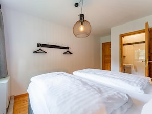 Une chambre avec deux lits, un luminaire suspendu, un porte-manteau et une porte en bois donnant accès à une salle de bain.