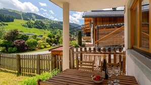 A wooden table with a bowl of fruit and a wine glass, a wooden fence, and a view of a green valley and mountains.