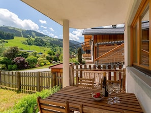 Ein Holztisch mit einer Obstschale und einem Weinglas, ein Holzzaun und ein Blick auf ein grünes Tal und Berge.