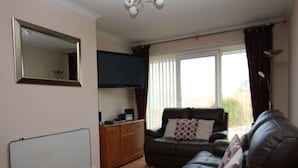 Ein Wohnzimmer mit braunem Ledersofa, einem weißen Heizkörper, einem am Wand montierten Flachbildfernseher und einem fenster mit Vorhängen.