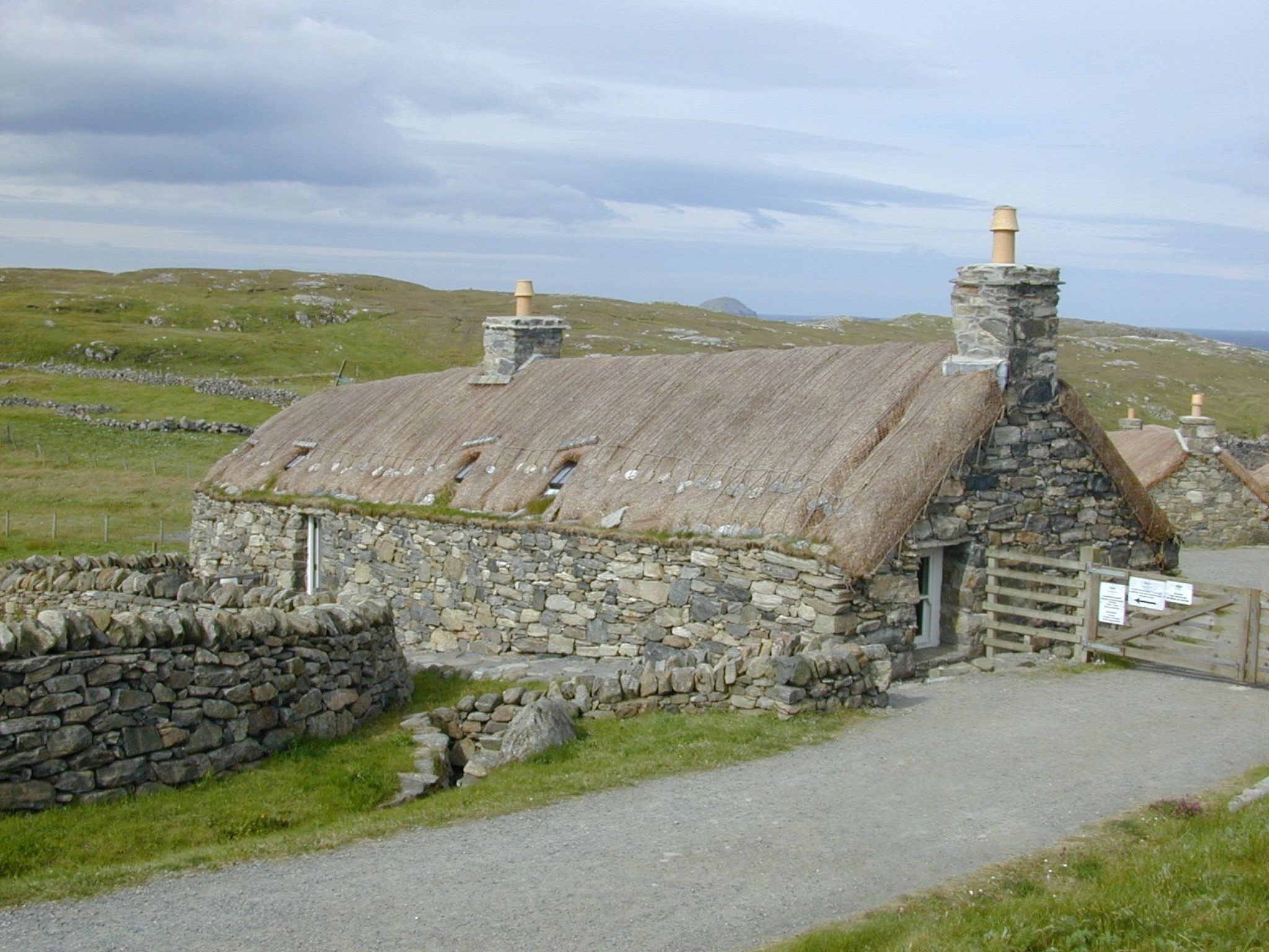 A stone building with a thatched roof, a stone wall, and a gravel path leading to it.