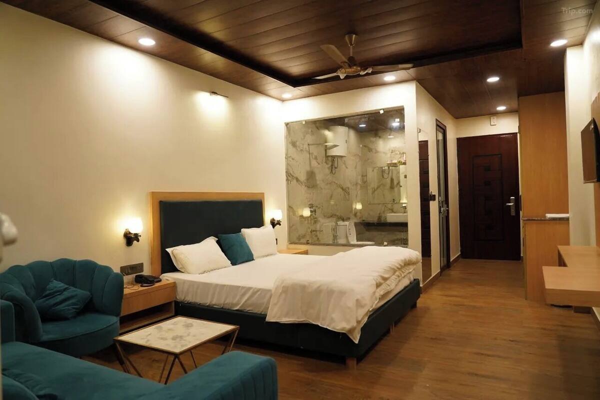 A modern hotel room with a bed, a seating area, a bathroom with a glass shower, and a wooden ceiling.