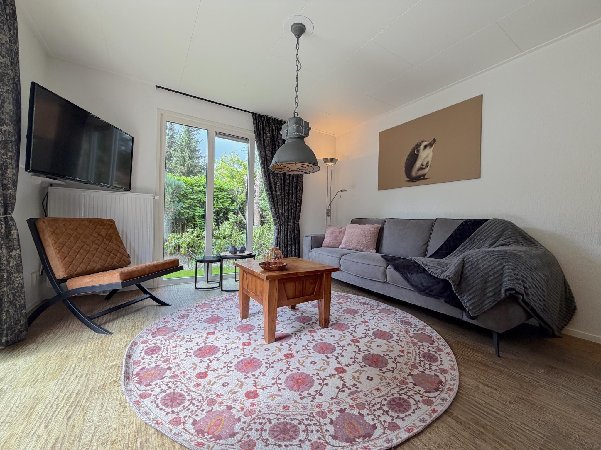 Ein Wohnzimmer mit einer Couch, einem Sessel, einem Couchtisch, einem Fernseher und einem großen Fenster mit Blick ins Grüne.