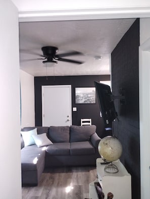 Ein Wohnzimmer mit einem grauen Ecksofa, einem Deckenventilator, einem Globus auf einem Stativ und einem schwarzen Fernseher an der Wand.