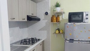Una cucina compatta con piano cottura a gas, forno a microonde e frigorifero, caratterizzata da mobili in legno chiaro e zoccolo paraschizzi rivestito in piastrelle.