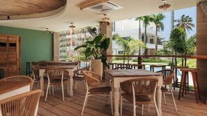 An outdoor dining area with wooden tables and chairs, a potted plant, and a view of palm trees and buildings. - 2BR/2.5BA Beachfront Condo | Sol de Arena B2 (Las Terrenas)