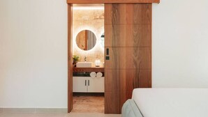 A modern bathroom with a wooden door, a round mirror, a sink, and cabinets. - 2BR/2.5BA Beachfront Condo | Sol de Arena B2 (Las Terrenas)
