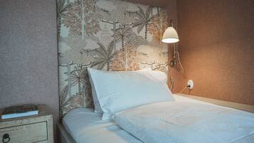 Una cama bien hecha con sábanas blancas, cabecera con estampado floral, mesita de noche con un libro y una lámpara fijada a la pared.