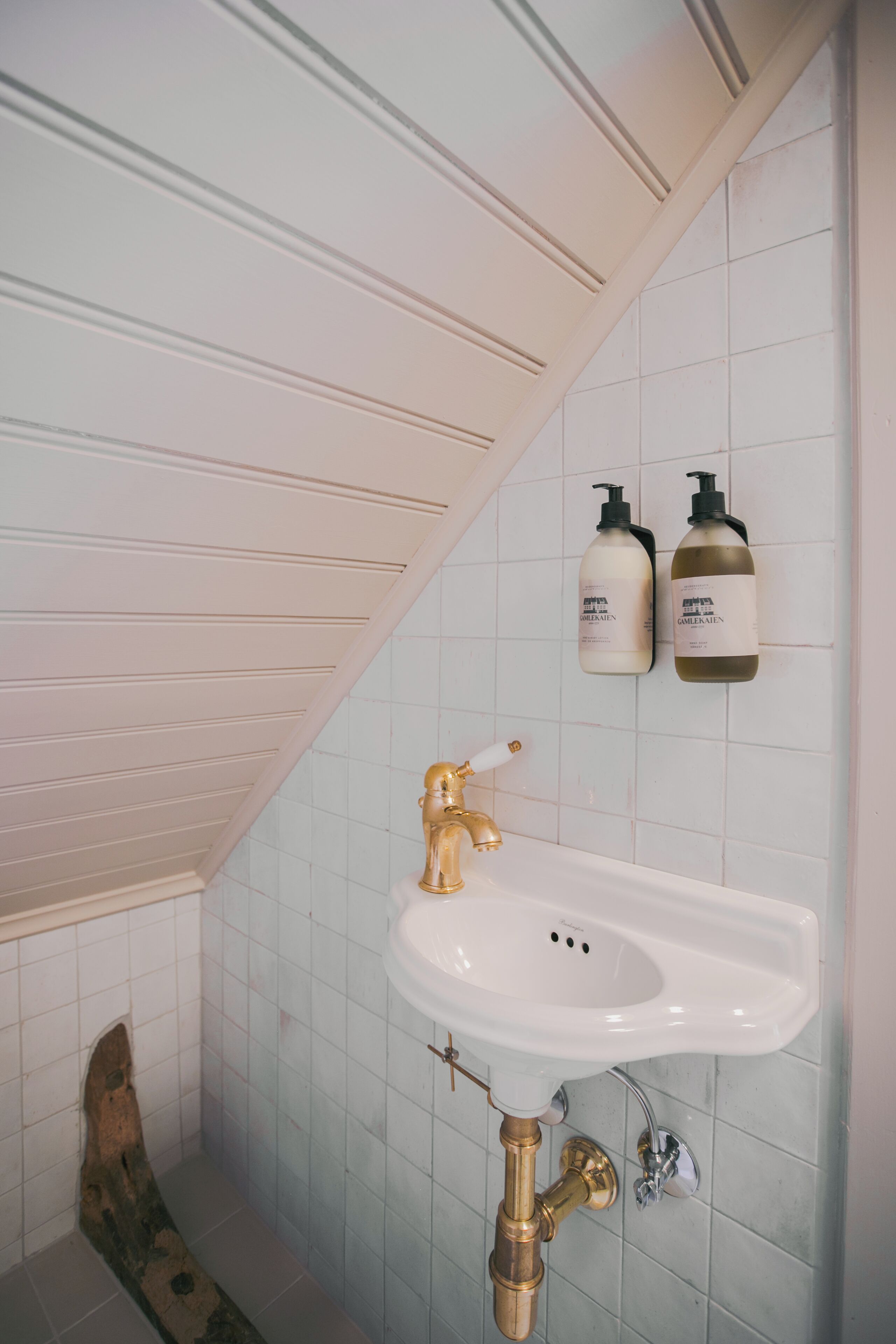 Un baño con lavamanos blanco, grifo dorado y dos botellas de jabón líquido en la pared.