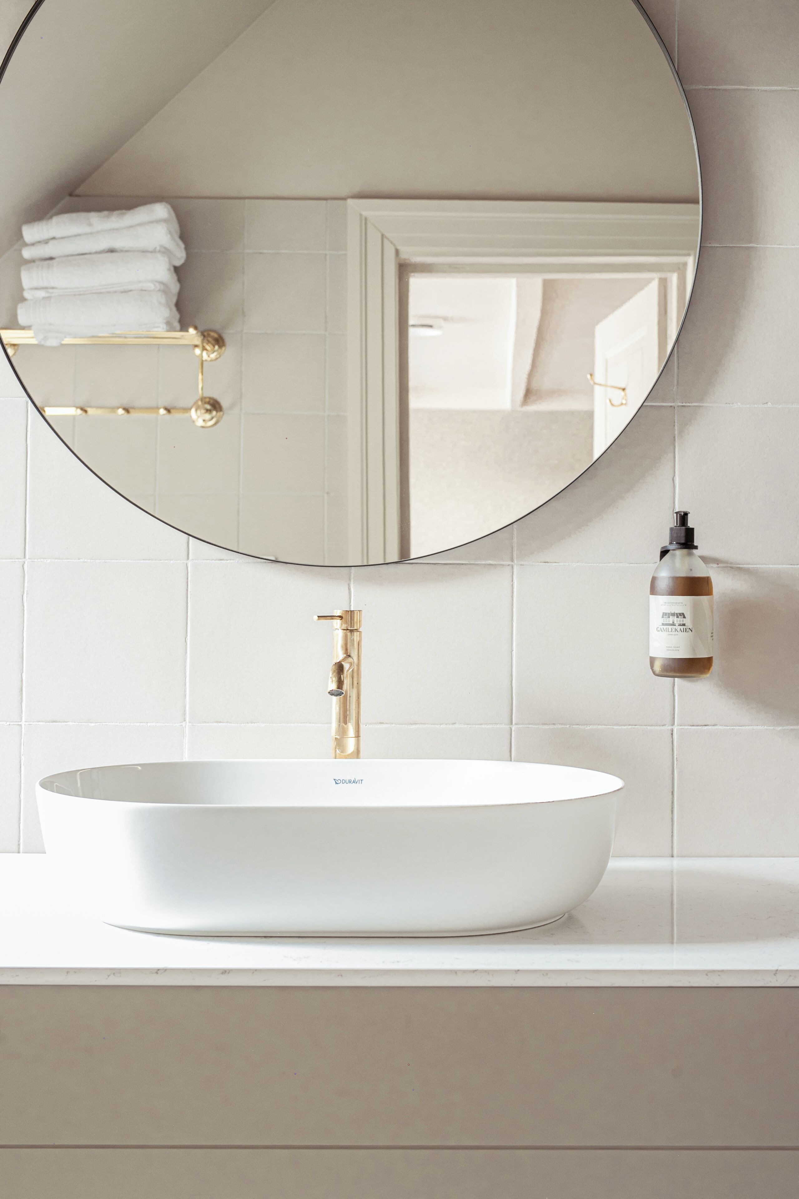 Un baño con un lavamanos blanco, un grifo dorado y un espejo redondo.