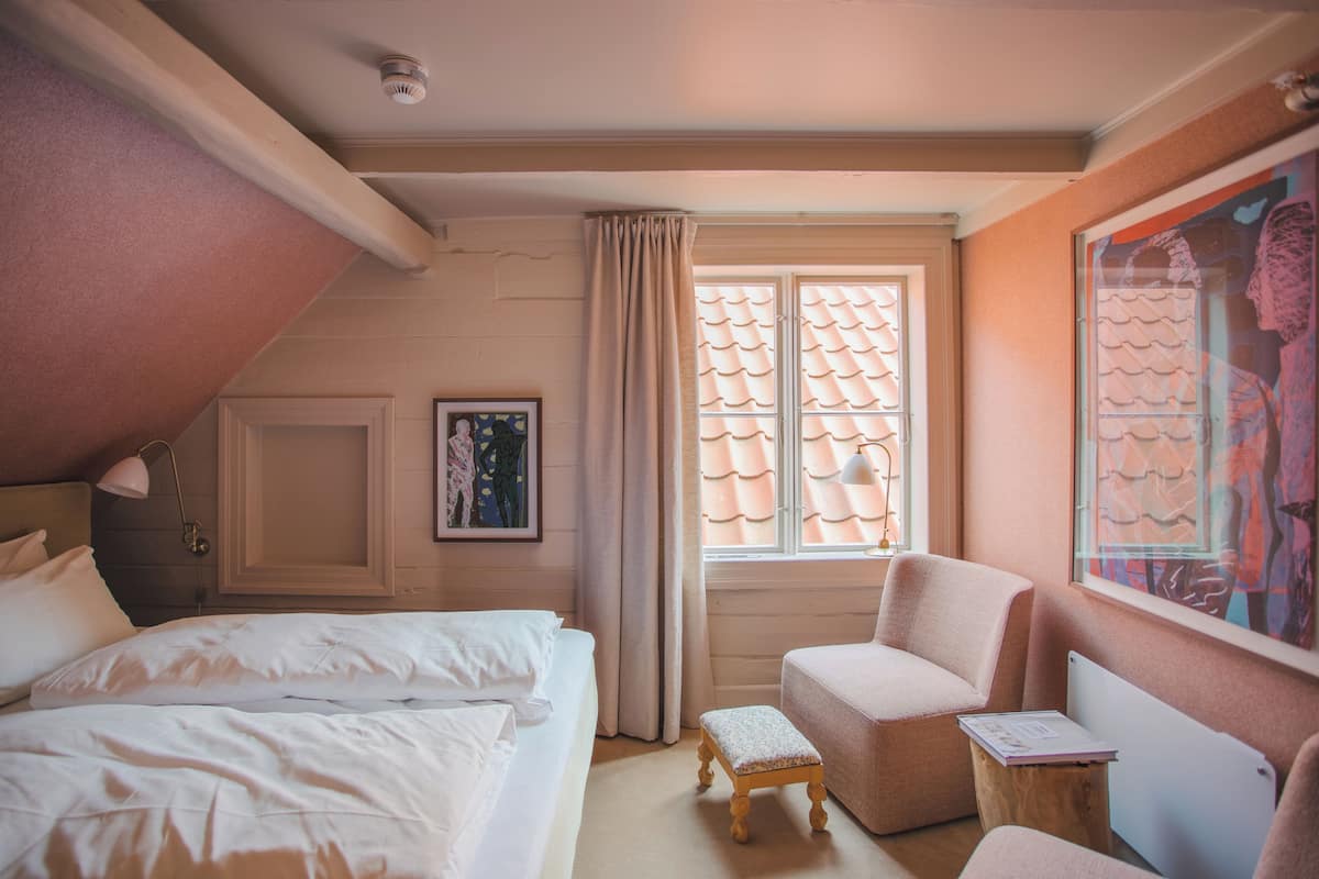 Een slaapkamer met een bed, een stoel, een klein tafeltje, een raam met gordijnen en een schilderij aan de muur.