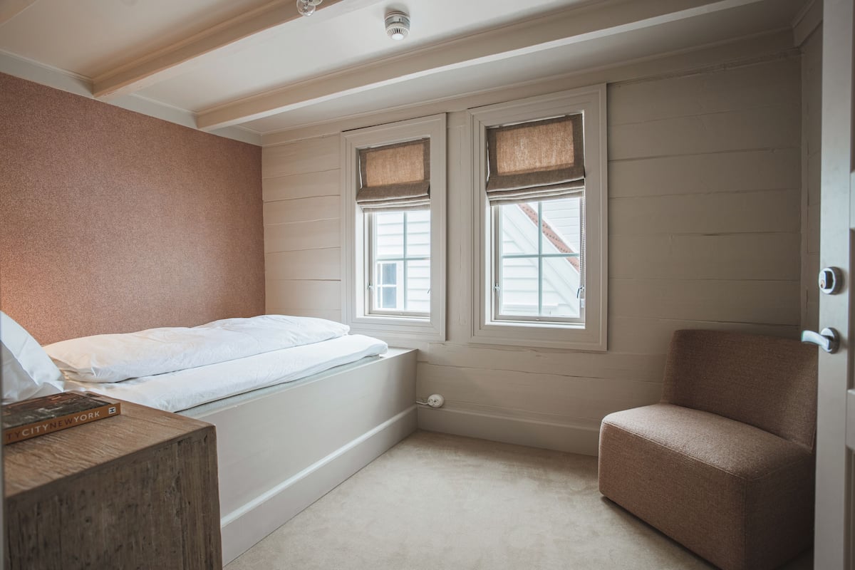 Een slaapkamer met een bed, een houten nachtkastje, een bruine stoel en twee ramen met luiken.