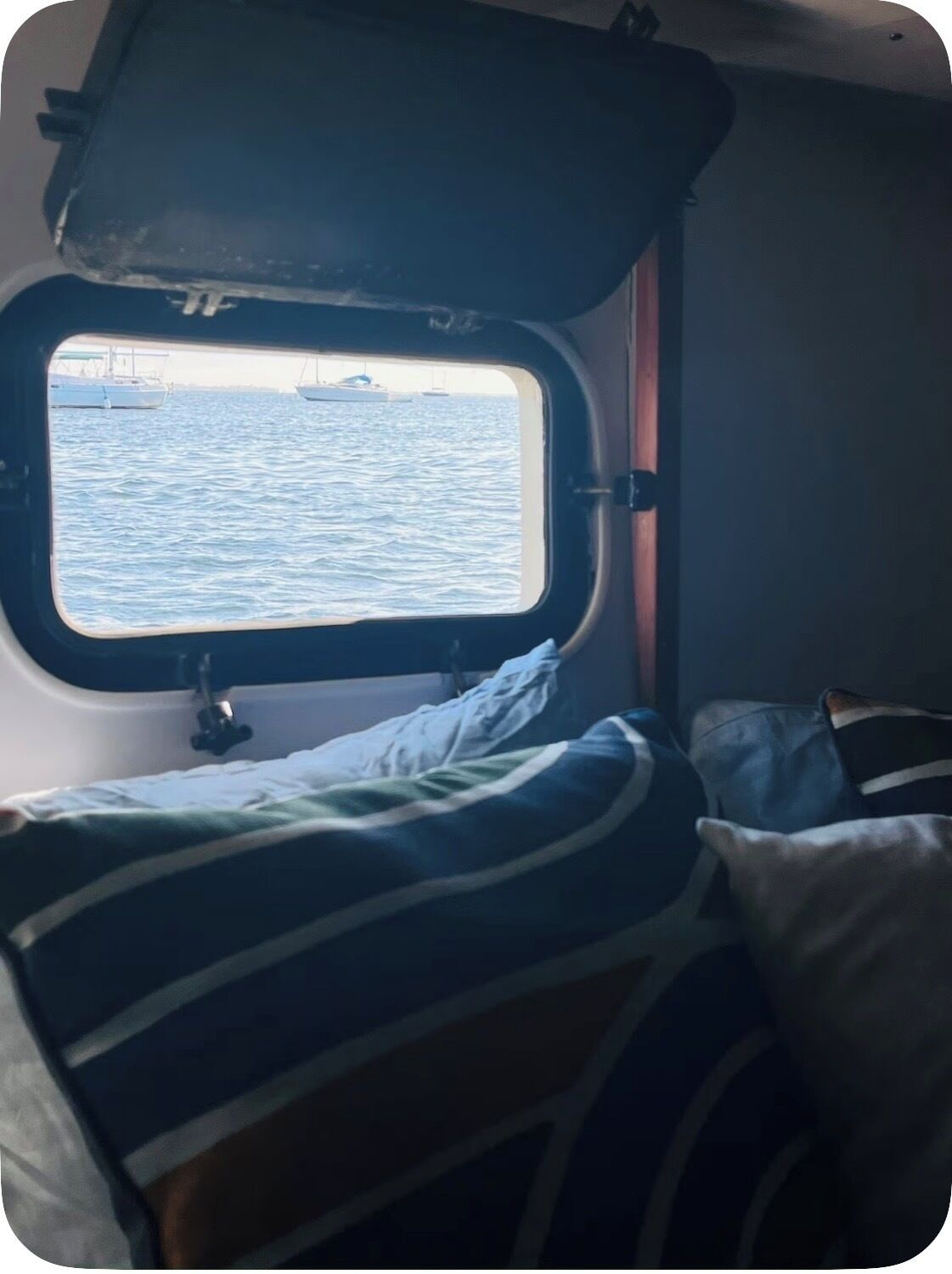 A small cabin with a window overlooking the sea, a bed with striped bedding, and a small table.