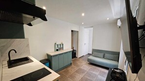 Een compacte keuken met een spoelbak, fornuis en een klein aanrecht. Er ligt een lichtgekleurde betegelde vloer en in de aangrenzende woonkamer staat een bank.