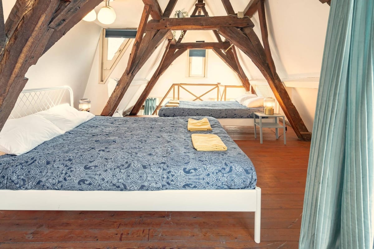 A bedroom with a wooden ceiling, a bed with a patterned bedspread, a bedside table with a lamp, and a window.