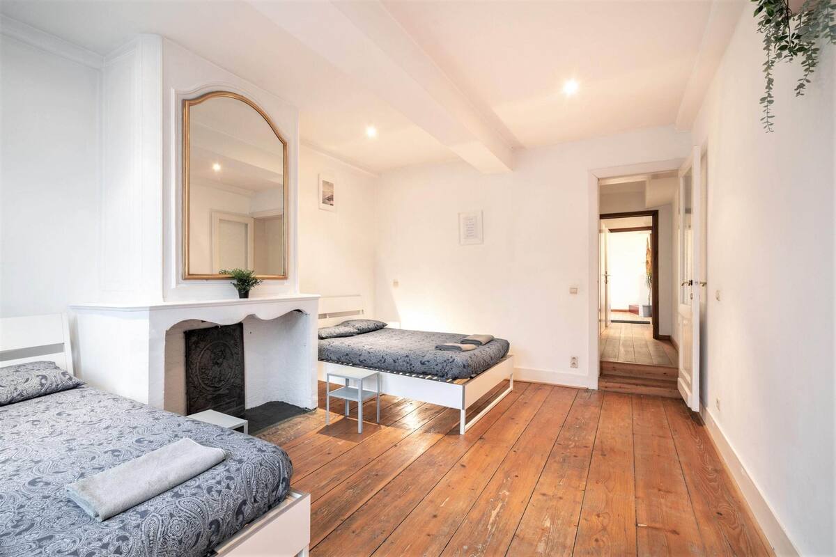 A bedroom with two beds, a mirror, a fireplace, and a hallway leading to another room.
