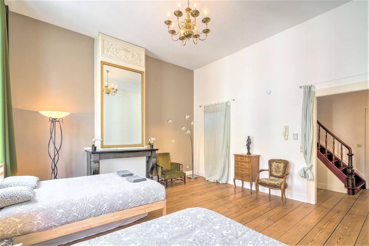 A bedroom with two beds, a large mirror, a chandelier, a staircase, and a green armchair.