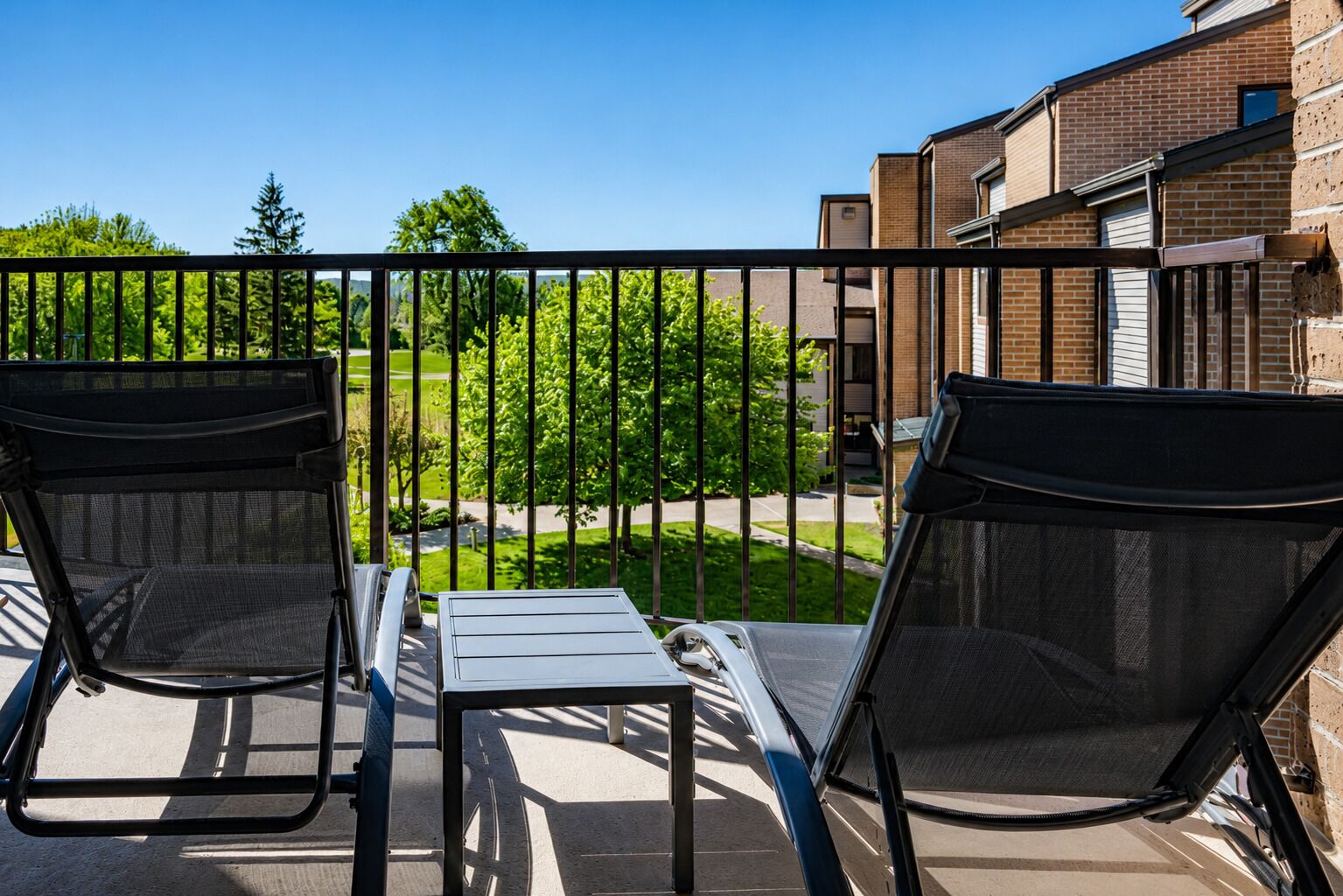 A balcony with two chairs and a small table, overlooking a green lawn and a brick building.