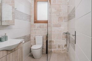 A bathroom with a stone wall, a glass shower enclosure, a white toilet, and a sink with a mirror. - Guince House by All About Paxos (Paxos)