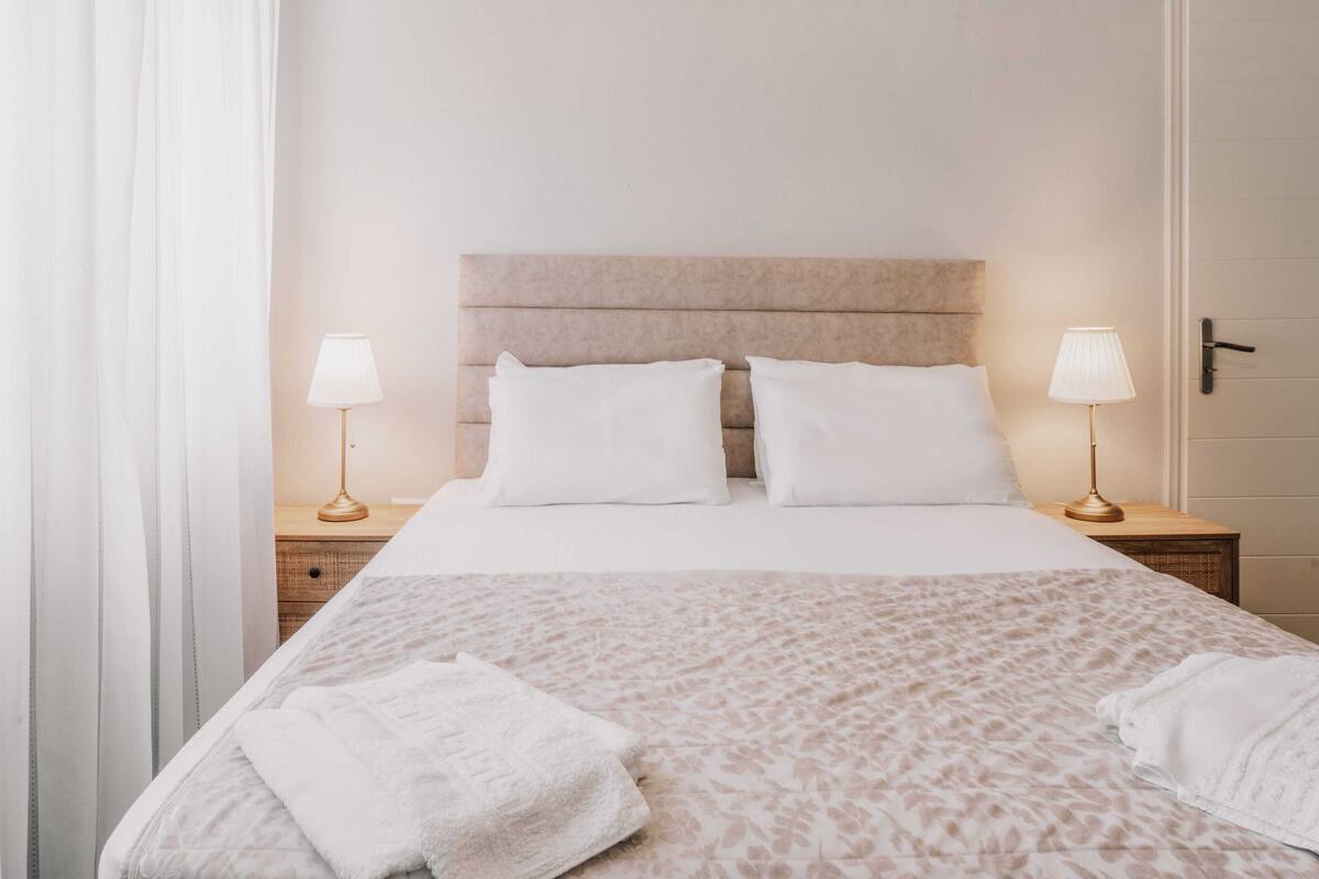 Quarto de hotel com uma cama, duas luminárias de cabeceira, uma mesinha de cabeceira e uma porta.