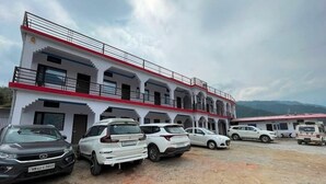 A multi-story building with a red and white facade, parked cars, and a mountainous backdrop. - rk tourist loge (Ukhimath)