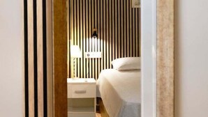 Ein schmaler Flur, der zu einem Schlafzimmer mit einem Bett, einem Nachttisch, einer Lampe und einem weißen, flauschigen Teppich führt.