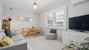 Un salon moderne comprenant un canapé, un téléviseur, une table à manger, des chaises et une fenêtre avec des stores.