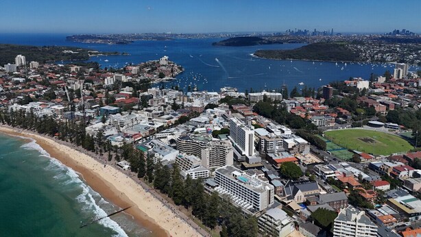 Vue aérienne d’une ville côtière avec une plage, des bâtiments et le profil d’une ville lointaine à l’horizon.