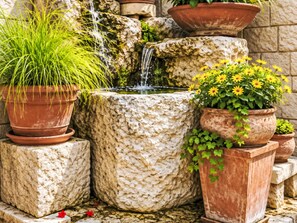 Ein Wasserbecken aus Stein mit einem Wasserfall, umgeben von Terrakotta-Pflanzgefäßen mit verschiedenen Pflanzen.