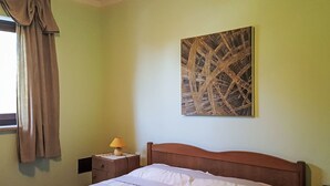 Ein Schlafzimmer mit einem Bett, einem Nachttisch, einer Lampe, einem Fenster mit Vorhängen und einem an der Wand hängenden abstrakten Gemälde.