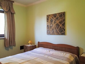 Ein Schlafzimmer mit einem Bett, einem Nachttisch, einer Lampe, einem Fenster mit Vorhängen und einem an der Wand hängenden abstrakten Gemälde.