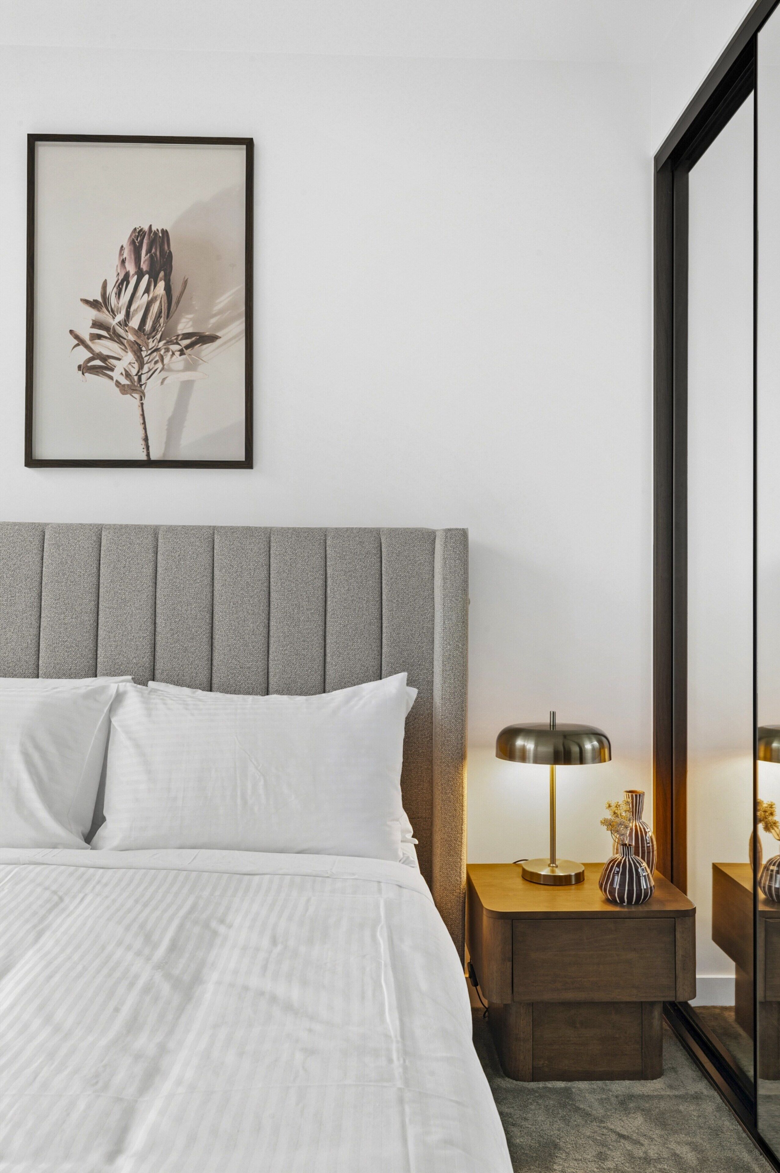 Ein ordentlich bezogenes Bett mit weißen Leinen, ein grauer möbelpolsterter Kopfteil, ein Nachttisch mit Lampe und ein botanisches Bild in einem Rahmen an der Wand.