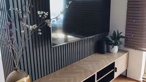 Un salon moderne avec un téléviseur à écran plat fixé au mur, un mur orné de lambris verticaux, un meuble de rangement en bois intégrant des espaces de stockage, un vase avec des branches et une plante en pot.